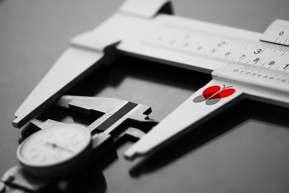Vernier calipers photographed close in high-contrast black and white, jaws open across a scale, with the red Visova butterfly mark sited on the sliding scale as the only color in the frame.