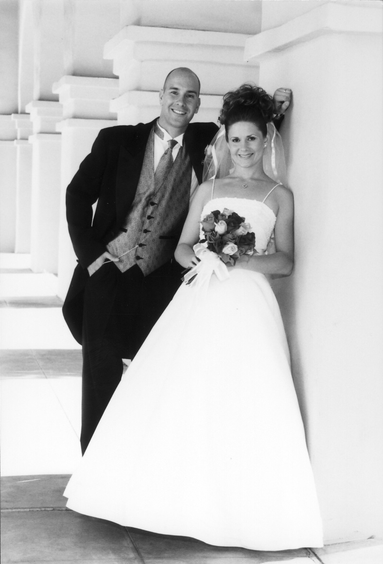 A black-and-white wedding portrait: the couple stand full-length against white columns, the bride in a full ball gown holding a bouquet, the groom in morning dress.